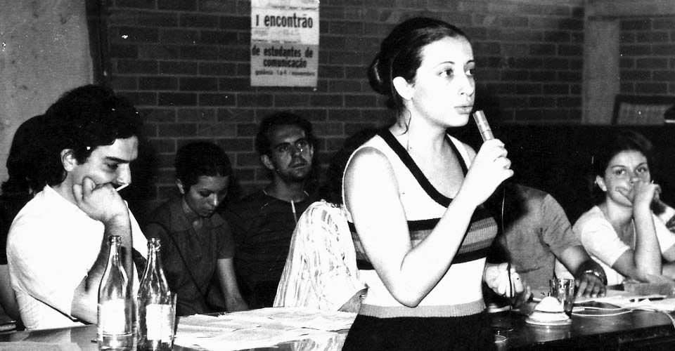 I ENECOM, Goiânia, 1972. Neuza Fiorda Chacha fala ao microfone. Ao fundo, o cartaz do "Encontrão (como era chamado este I Encontro), de autoria de Laerte Coutinho (contribuição de Neuza Fiorda Chacha)