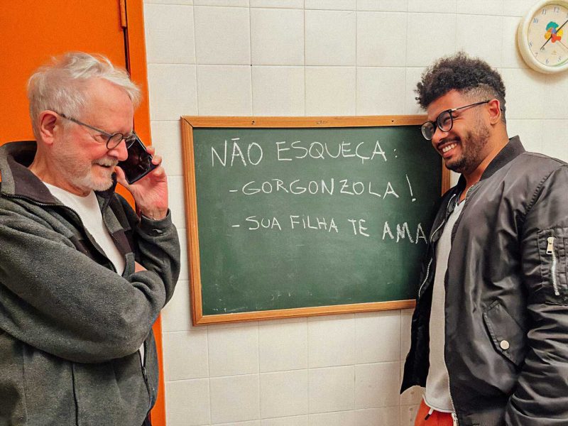 Jean-Claude e Diego na frente da lousa para recados, em foto tirada por Tata Amaral, em 2024, enquanto Jean-Claude falava com a filha, Lígia
